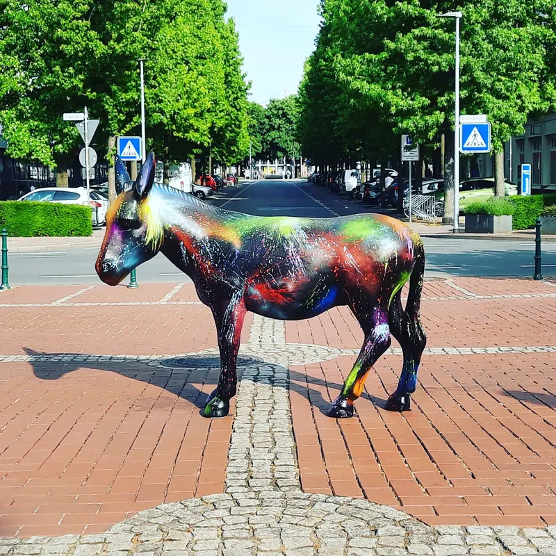 Schwarze Esel-Skulptur mit bunten Farbspritzern auf einem gepflasterten Platz, dahinter eine Allee