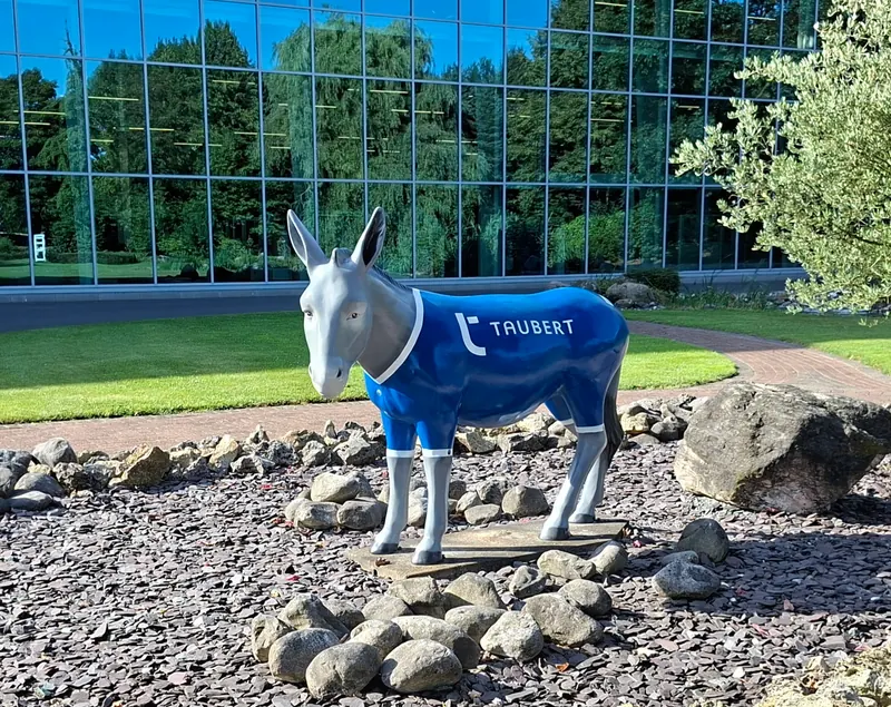 Graue Esel-Skulptur mit blauem Trikot und Firmenlogo auf Steinen vor einer Glasfassade
