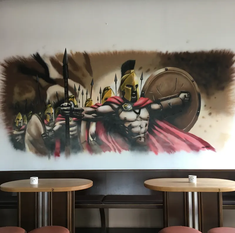 Wandbild mit spartanischen Kriegern in Rot und Braun, mit Speer und Schild, über Stehtischen in einem Gastraum
