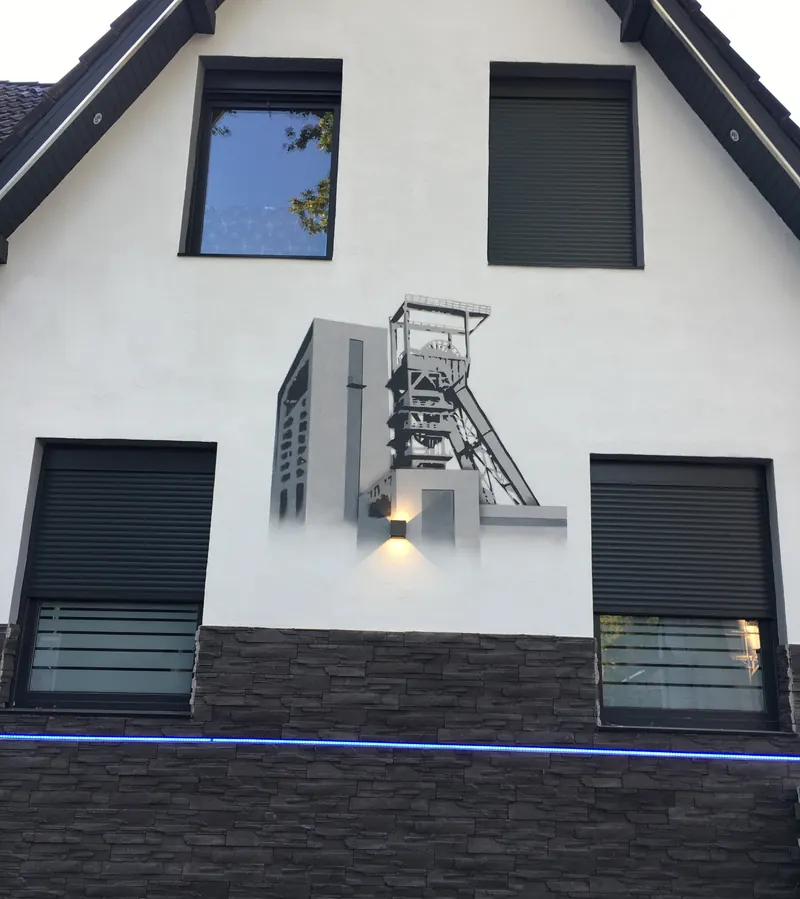 Weißer Hausgiebel mit gesprühtem Förderturm in Grautönen zwischen zwei Fenstern, darunter eine Außenleuchte und Steinverkleidung