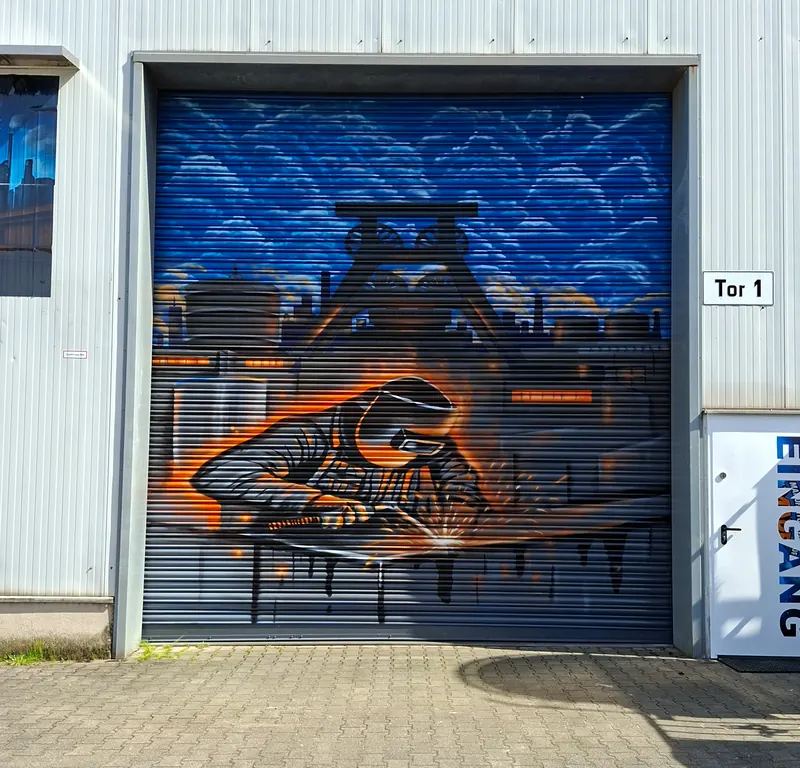 Rolltor mit Graffiti: Förderturm vor blauem Himmel, davor ein Schweißer mit Schutzmaske im orangefarbenen Funkenflug
