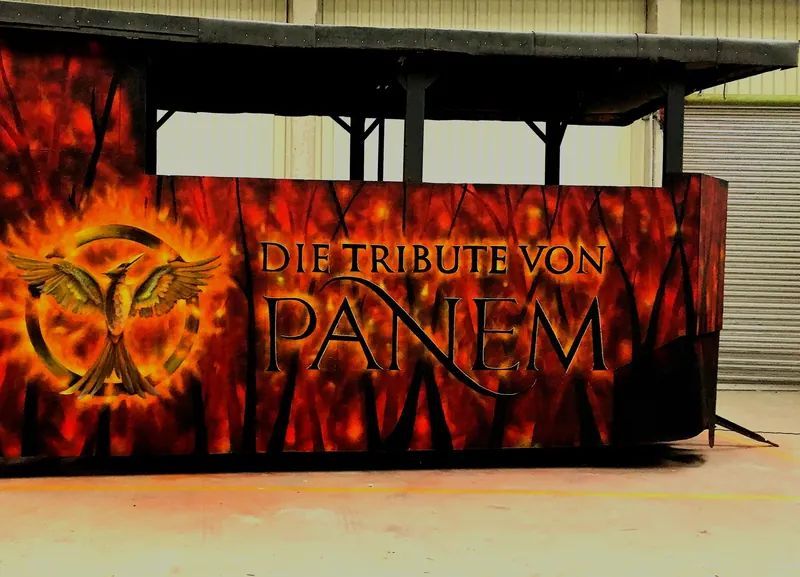 Stand mit Flammen-Graffiti in Rot und Orange, goldenem Vogel-Emblem und Schriftzug Die Tribute von Panem