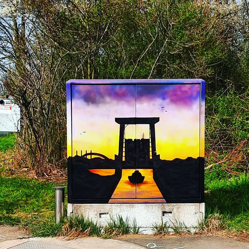 Stromkasten vor Büschen, bemalt mit lila-orangem Abendhimmel, Silhouette eines Tors am Wasser, Brückenbogen und einem Boot