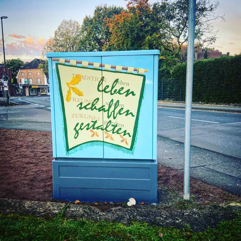 Hellblauer Stromkasten mit gemaltem Banner und dem Schriftzug leben schaffen gestalten, daneben Eichenblätter, im Hintergrund eine Straßenkreuzung