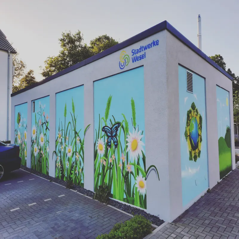 Trafostation mit Schriftzug Stadtwerke Wesel, die Wände bemalt mit hohen Gräsern, Margeriten und einem blauen Schmetterling, an der Seite eine Erdkugel im Blätterkranz