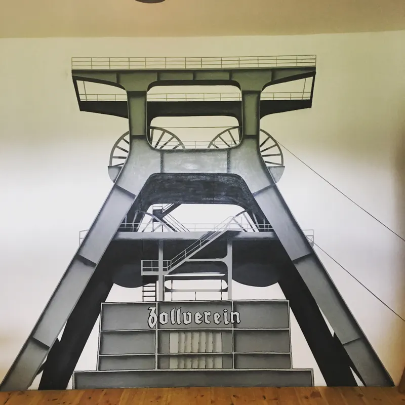Wandbild in Grautönen: Fördergerüst der Zeche Zollverein von unten gesehen, mit Schriftzug Zollverein